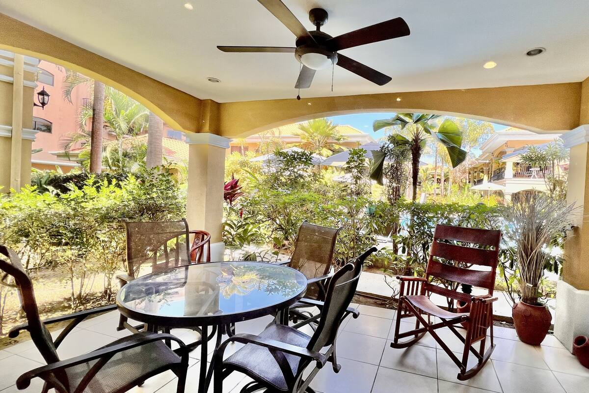 A covered patio with a round glass table and chairs, a ceiling fan, and potted plants.