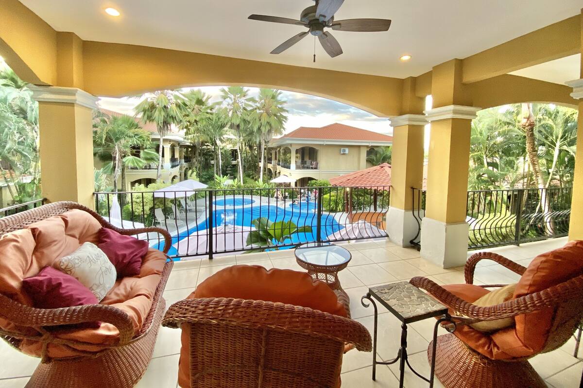 A covered outdoor area with wicker furniture, a pool, and a view of a building with a red roof.