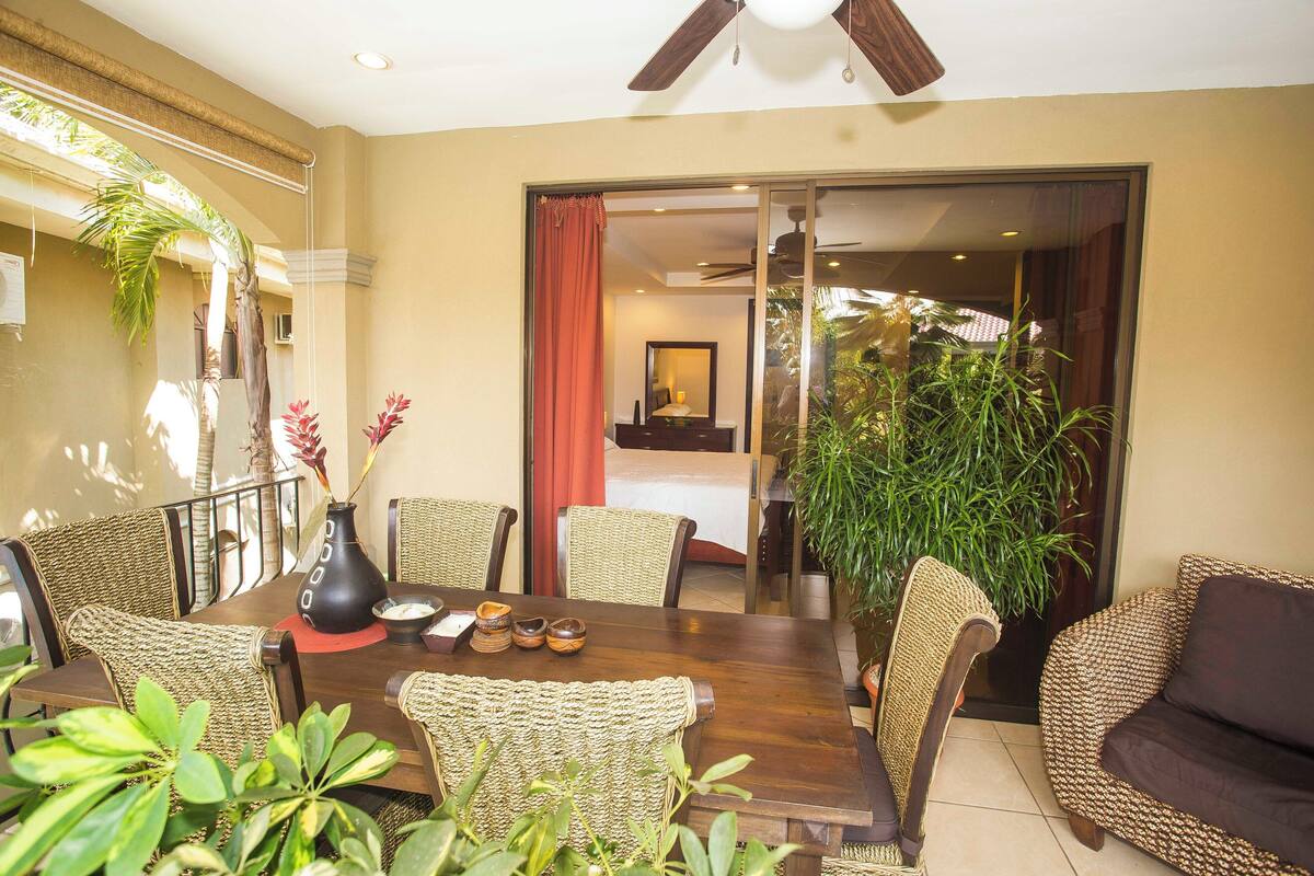 A dining area with a wooden table, wicker chairs, and a large potted plant.