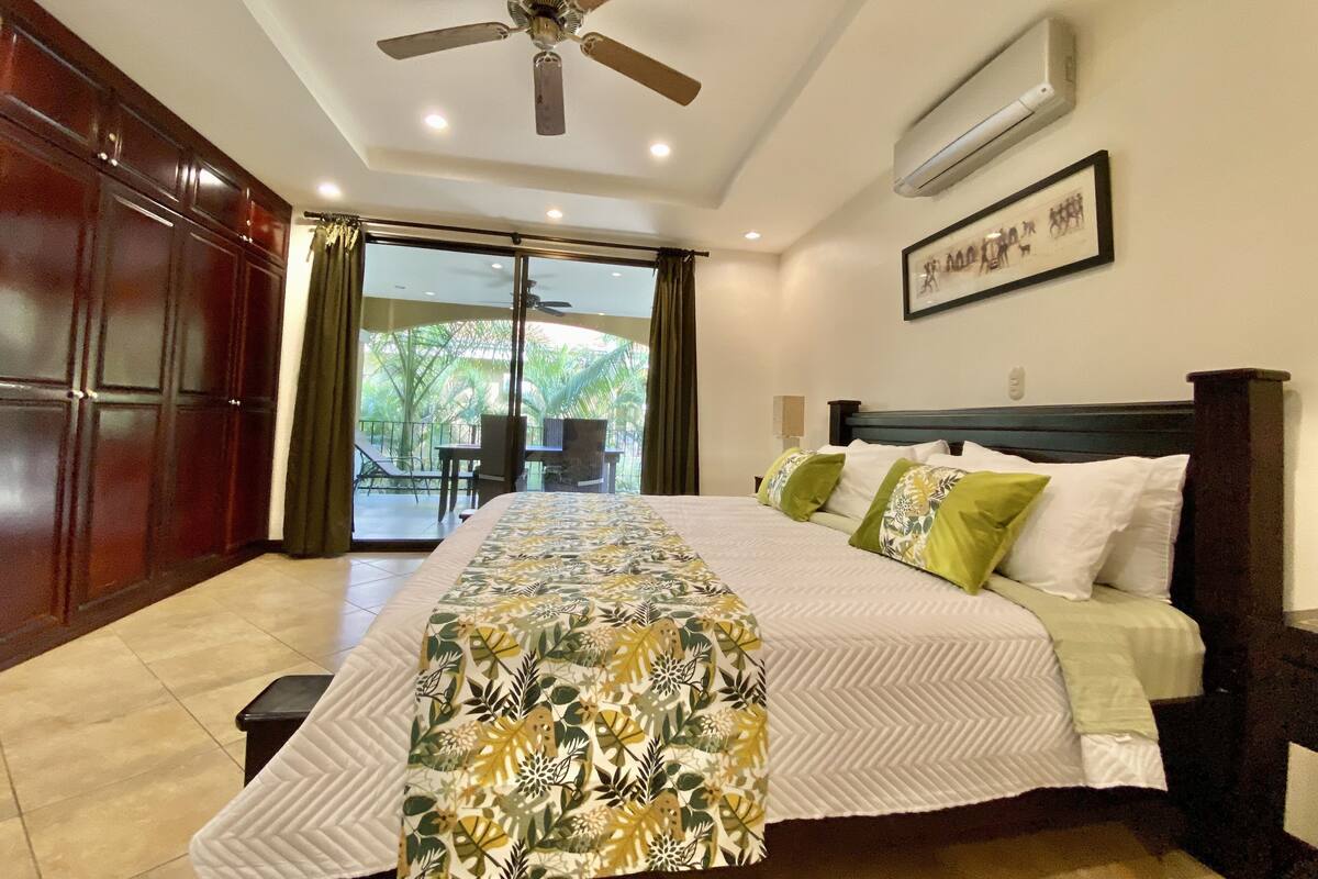 A bedroom with a bed, a ceiling fan, an air conditioning unit, a window with a view of palm trees, and a door leading to a balcony.