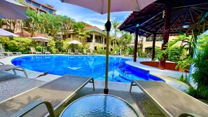 Una piscina con lettini e ombrelloni, immersa nel verde e affiancata da un edificio con tetto rosso.
