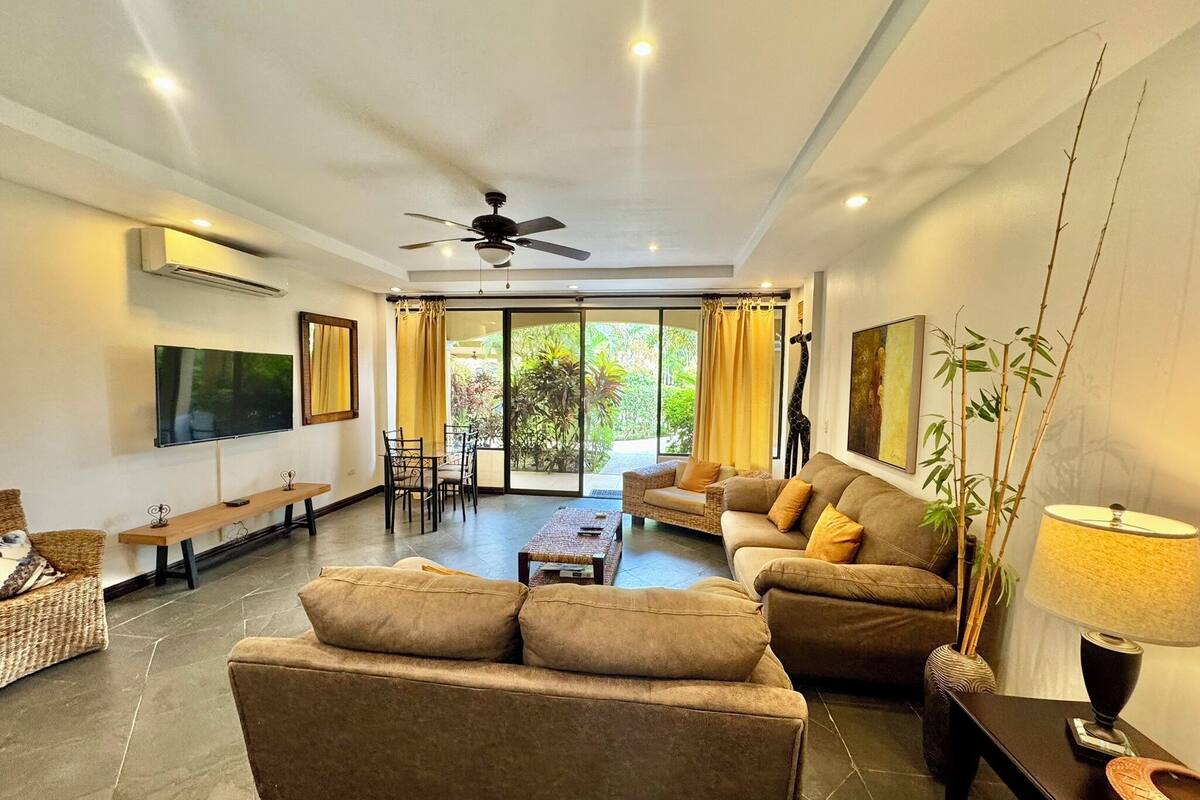 A spacious living area with a sofa, coffee table, and a TV mounted on the wall. There is a ceiling fan, a window with curtains, and a view of greenery outside.