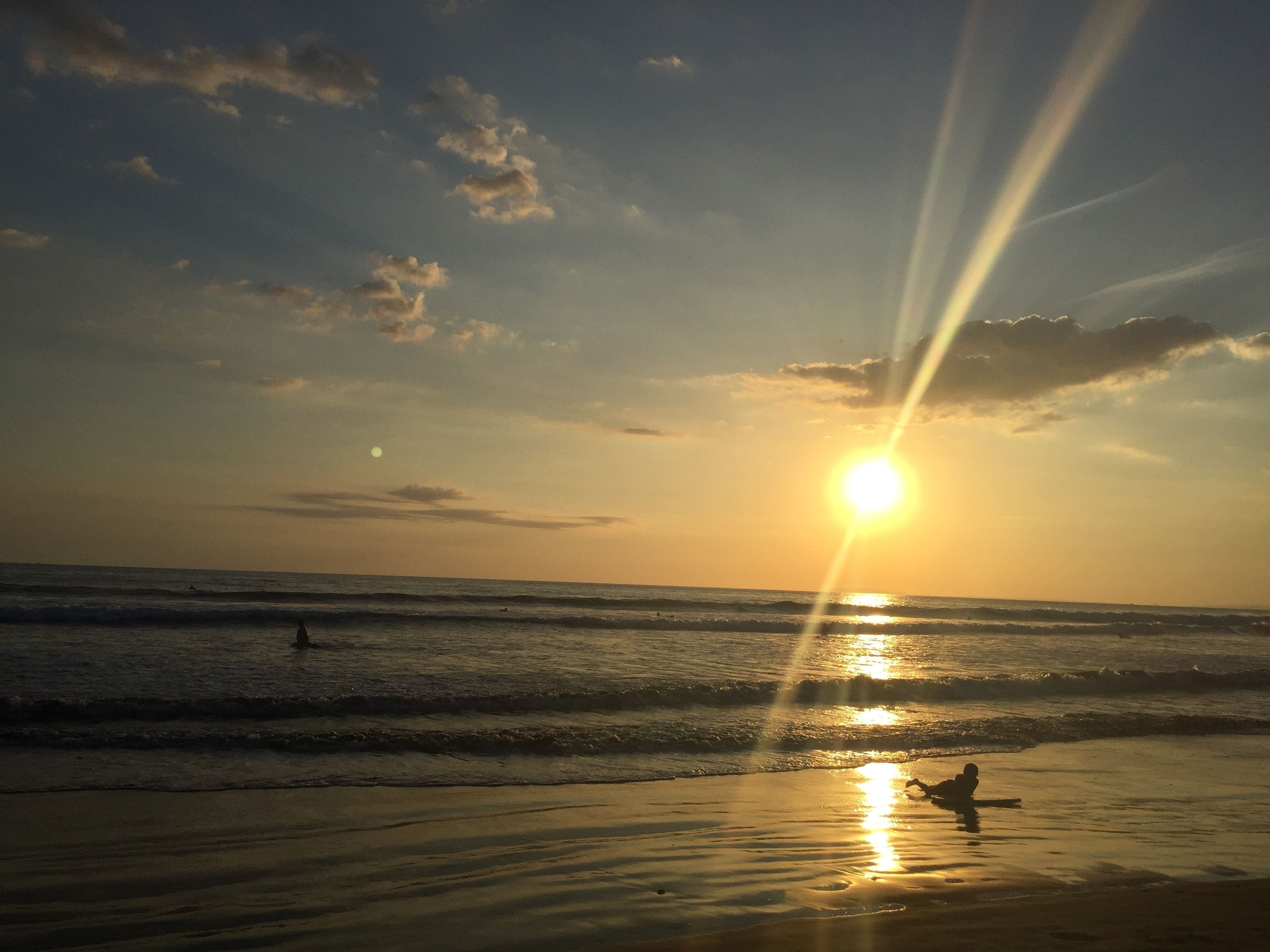 A beach scene with the sun setting over the ocean, waves gently lapping at the shore, and a person sitting on the sand.