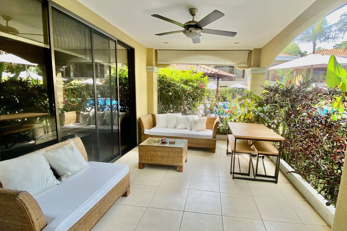 A balcony with a sofa, a table, and chairs. There is a ceiling fan and a view of the outside.