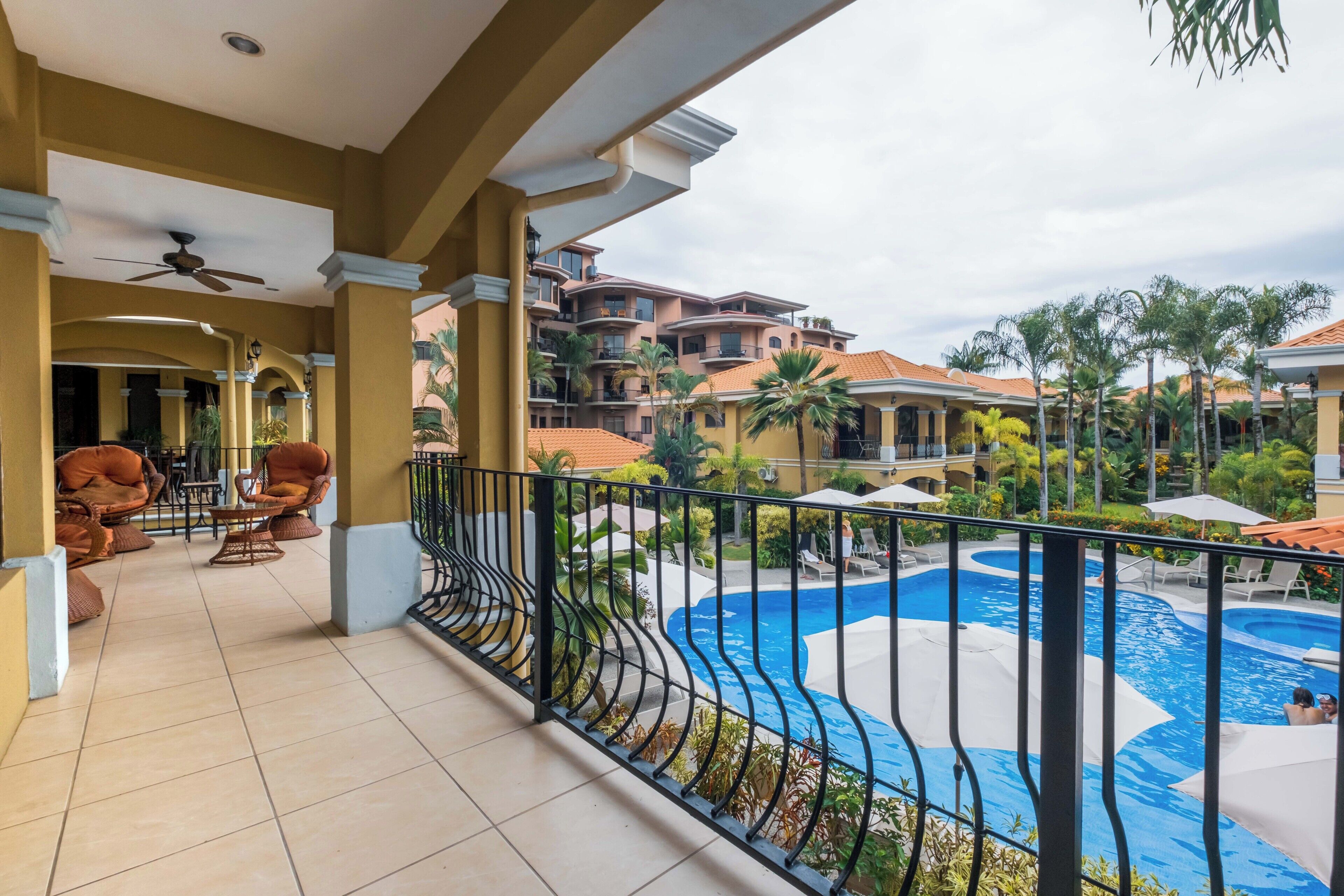 A balcony with a view of a swimming pool, lounge chairs, and a ceiling fan.
