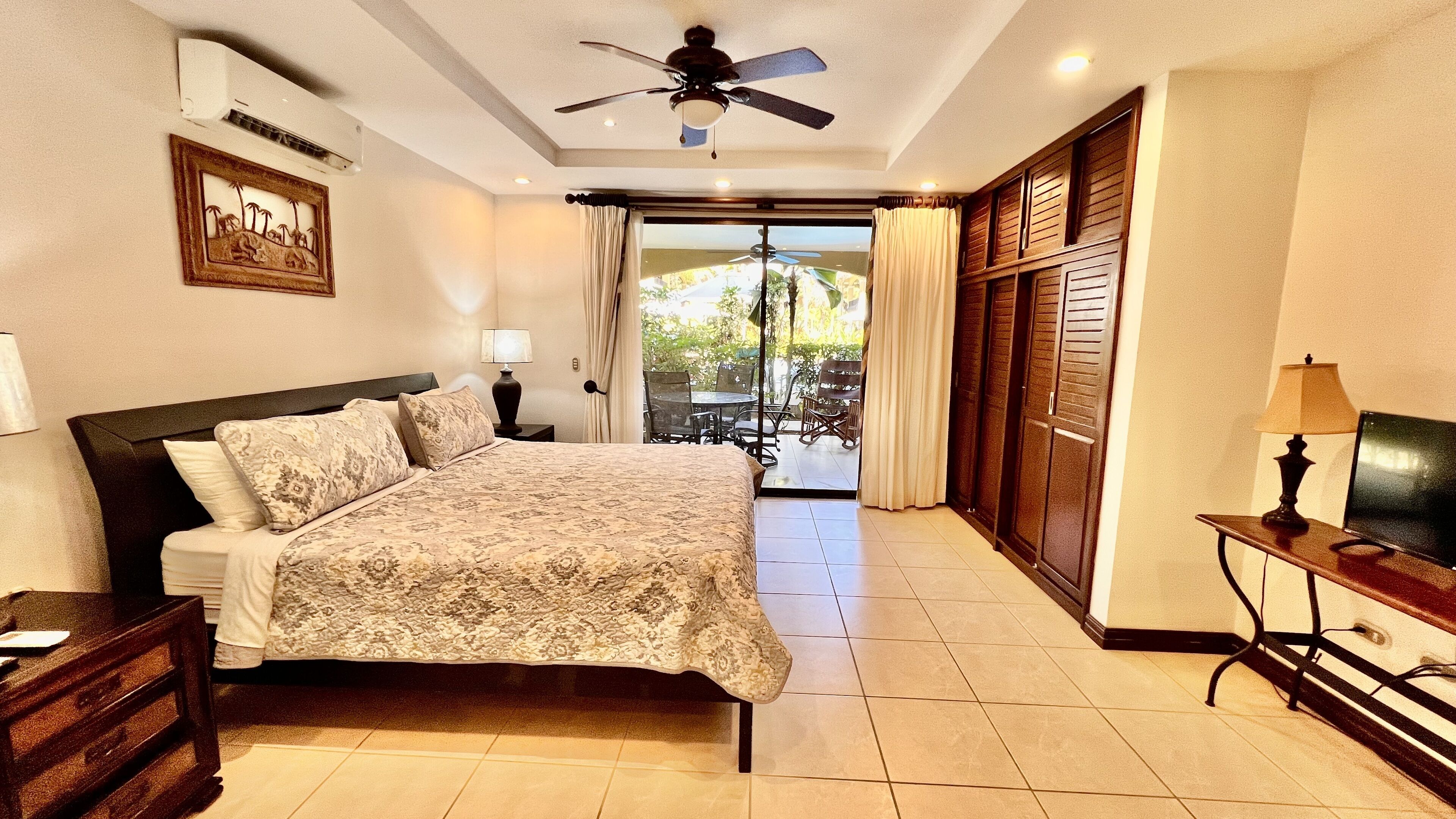 A bedroom with a bed, a ceiling fan, a nightstand, a lamp, a TV, and a view of the outside.