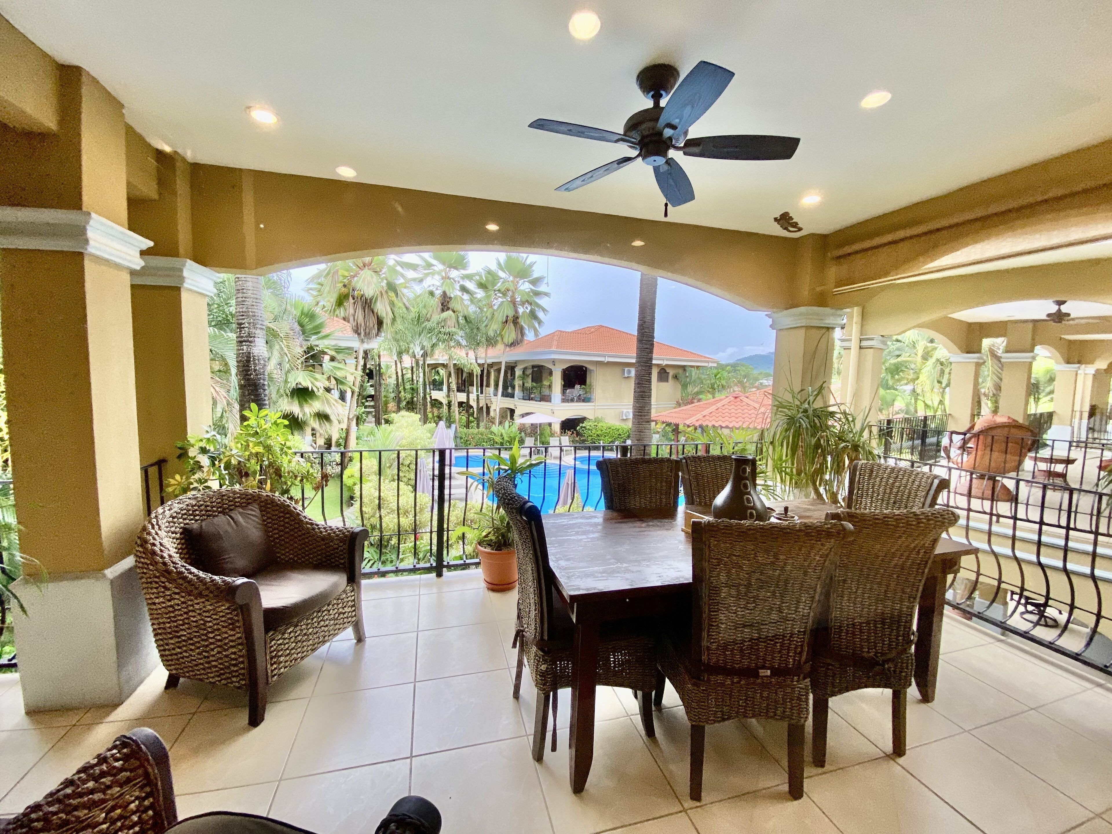 A covered outdoor area with wicker furniture, a ceiling fan, and a view of a pool and building in the background.