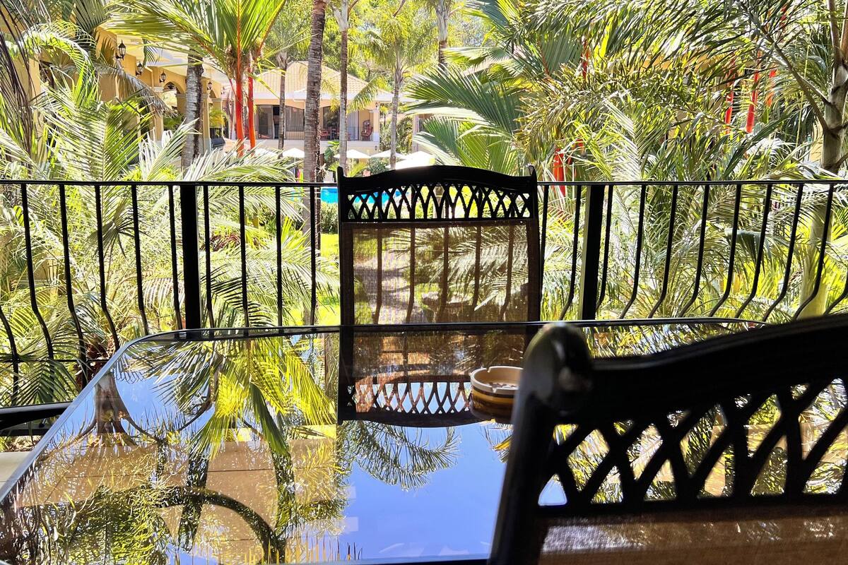 A balcony with a glass table and chairs, surrounded by a metal railing and lush tropical plants.