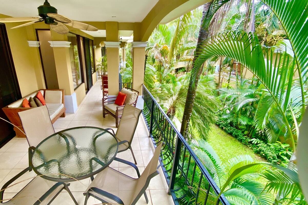 A balcony with a glass table and chairs, a sofa with cushions, and a view of lush greenery.
