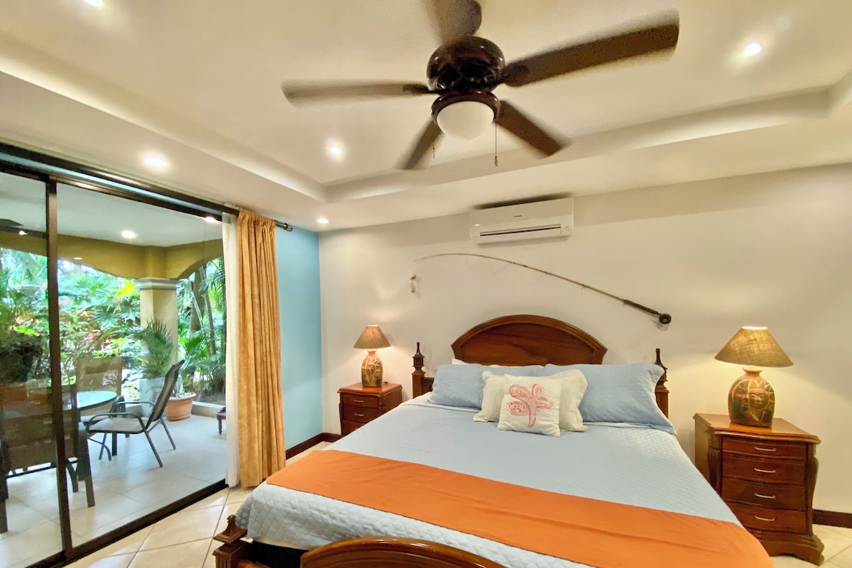 A bedroom with a bed, two bedside tables, a ceiling fan, and a view of a patio with plants.