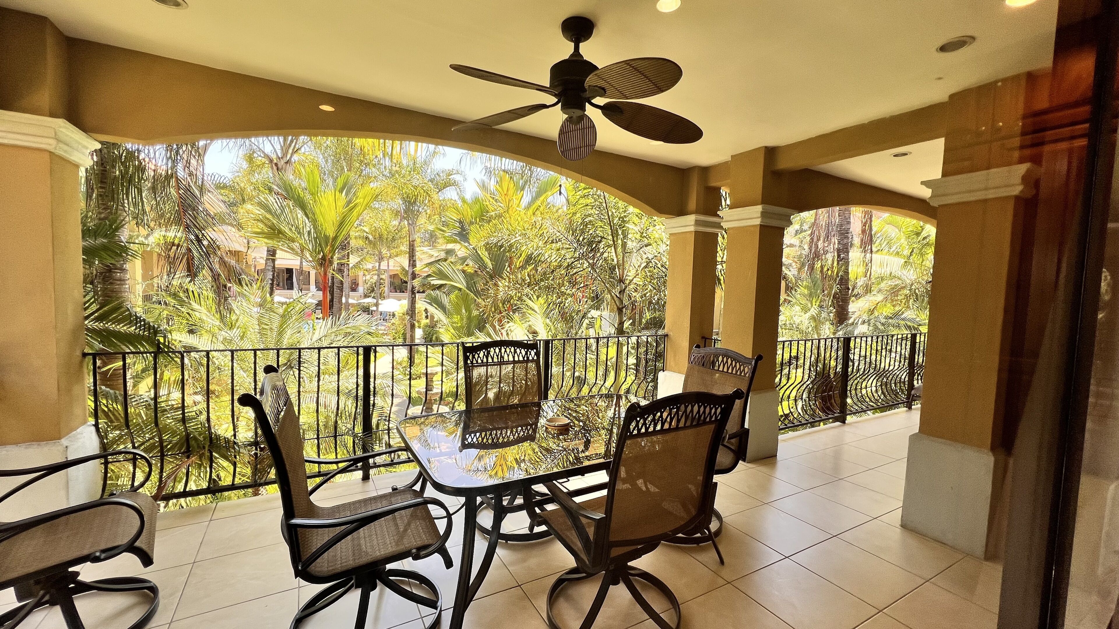 A covered outdoor seating area with a ceiling fan, surrounded by a metal railing, overlooking a lush garden with palm trees.