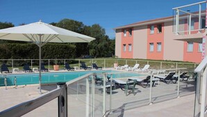 A pool area with lounge chairs and a large umbrella, a glass balustrade, and a building with a pink facade in the background.