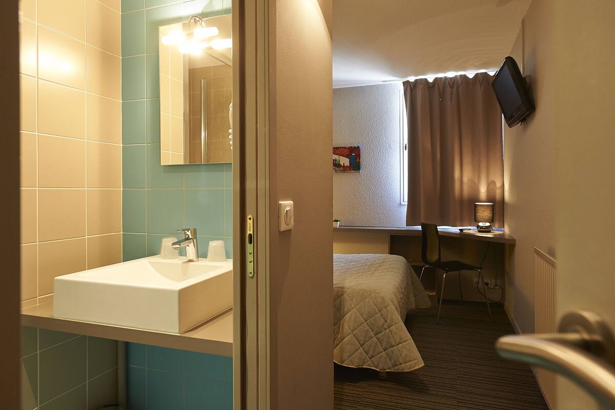 A hotel room with a bathroom featuring a sink and mirror, a bed with a quilted cover, a desk with a chair, and a television mounted on the wall.