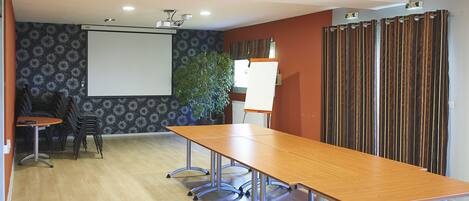 Un salón de conferencias con una larga mesa, sillas, una pantalla de proyector y una planta.