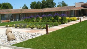 A building with a tiled roof, a garden with various plants and flowers, a paved walkway, and a grassy area.