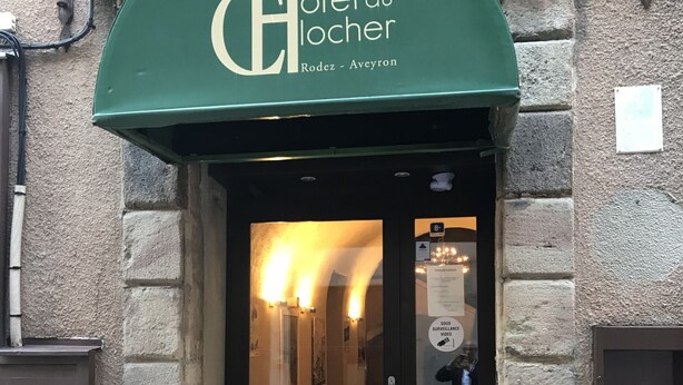 La entrada del Hotel du Locher en Rodez-Aveyron, con un toldo verde, una puerta de cristal y un pequeño espacio exterior con mesas y sillas.