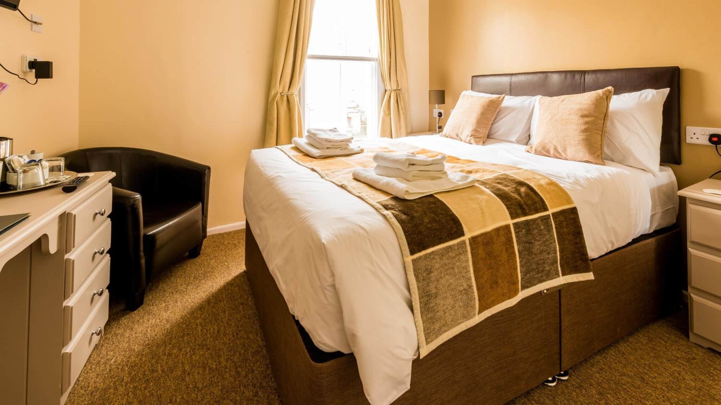 Una cama bien hecha con cabecera marrón, una mesita de noche con lámpara y un escritorio con silla.