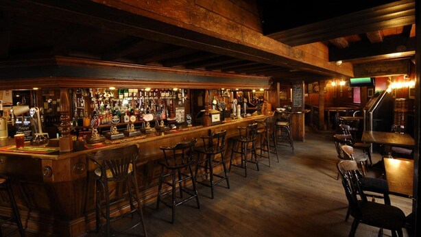 A bar area with wooden beams, a bar counter, and multiple stools. - The Longboat Inn (Penzance)