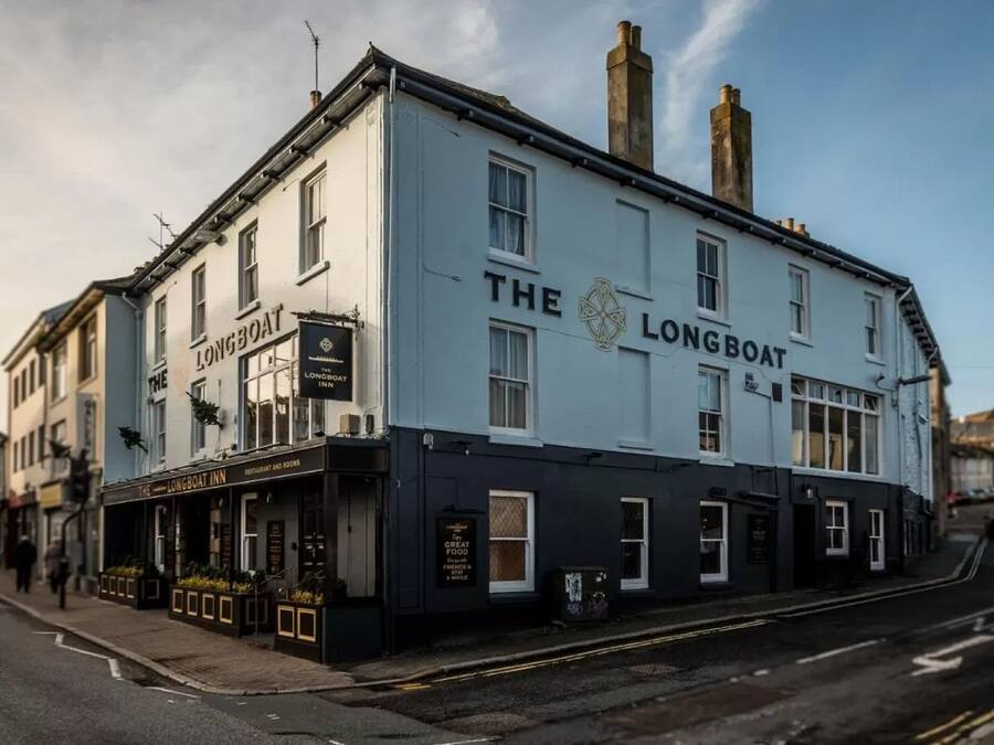 Un bâtiment à deux étages portant l’enseigne « The Longboat Inn ». Il se distingue par sa façade noire et blanche, ses nombreuses fenêtres et sa cheminée bien visible.