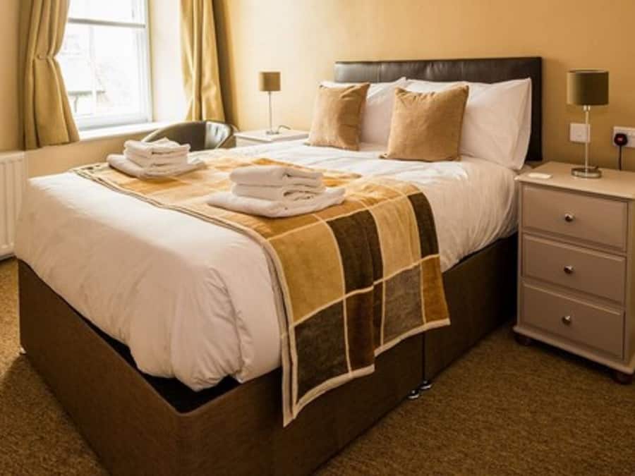 A neatly made bed with a brown headboard, a bedside table with a lamp, and a window with curtains.