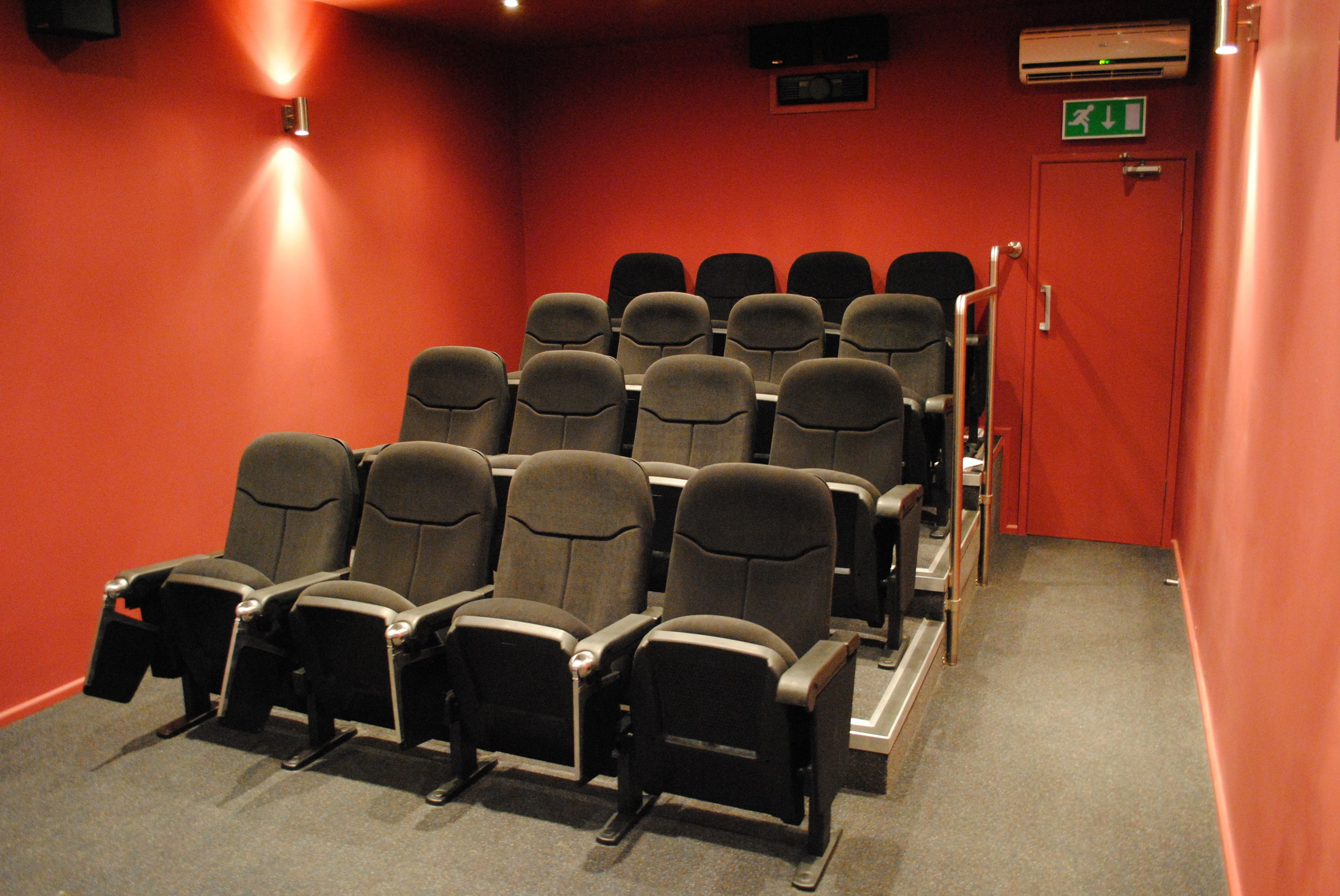 Una fila de asientos negros tipo sala de teatro en una habitación de paredes rojas, con un equipo de aire acondicionado y un letrero de salida de emergencia en la pared.
