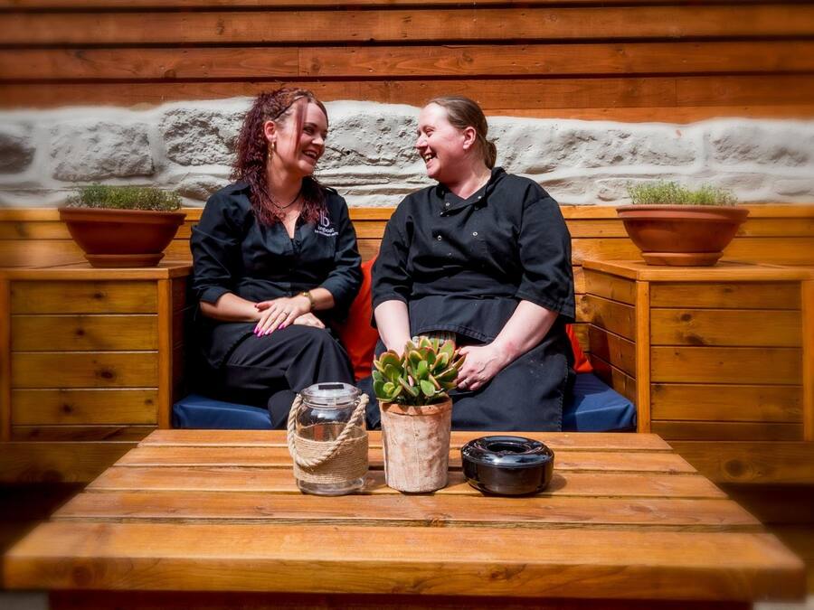 Two people seated on a bench with a wooden table in front of them, holding a plant and a small jar.
