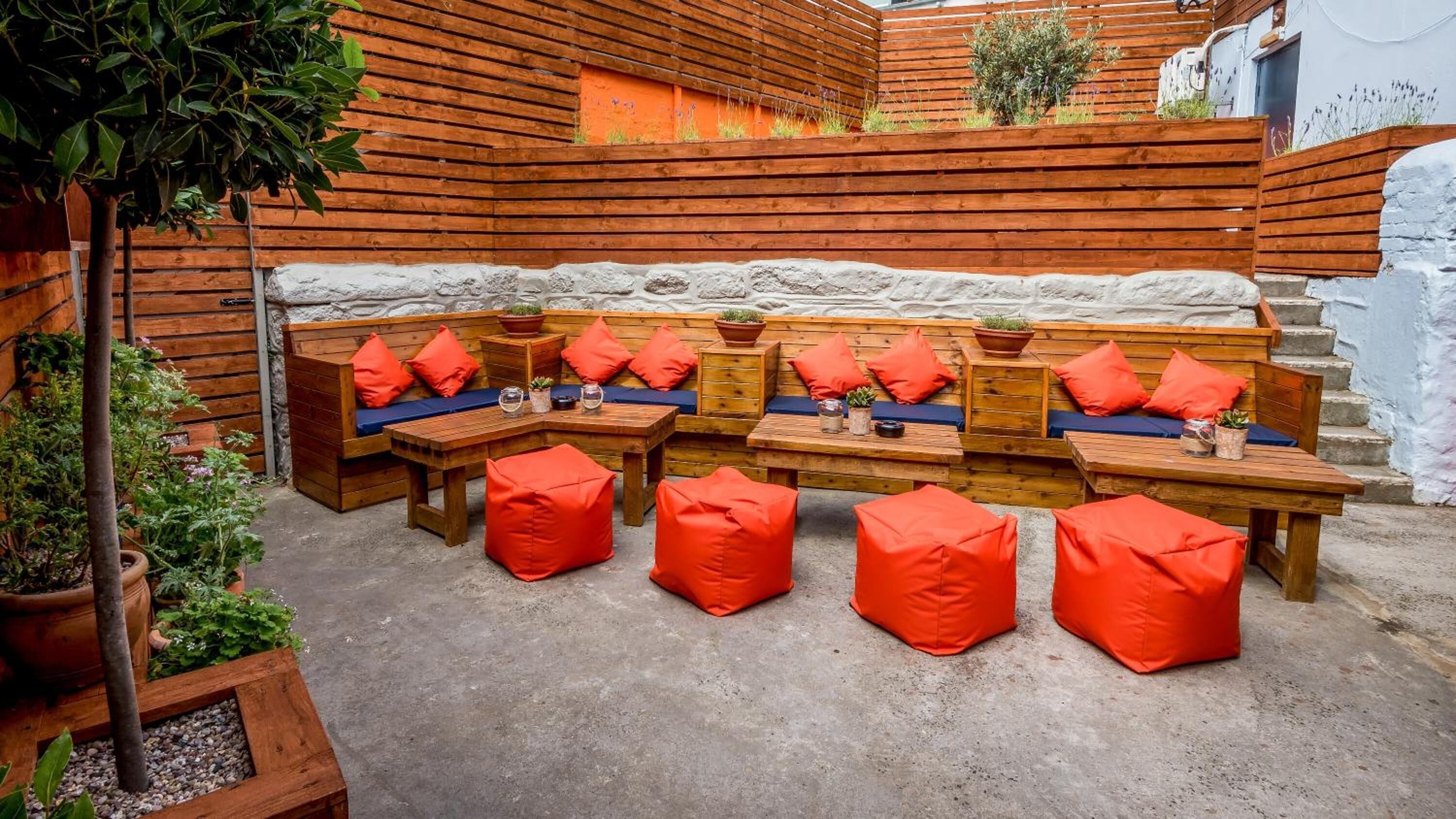 A wooden outdoor seating area with benches and tables, surrounded by plants and a stone wall.