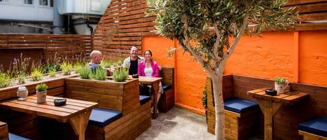A cozy outdoor seating area with wooden benches and tables, surrounded by plants and a tree.