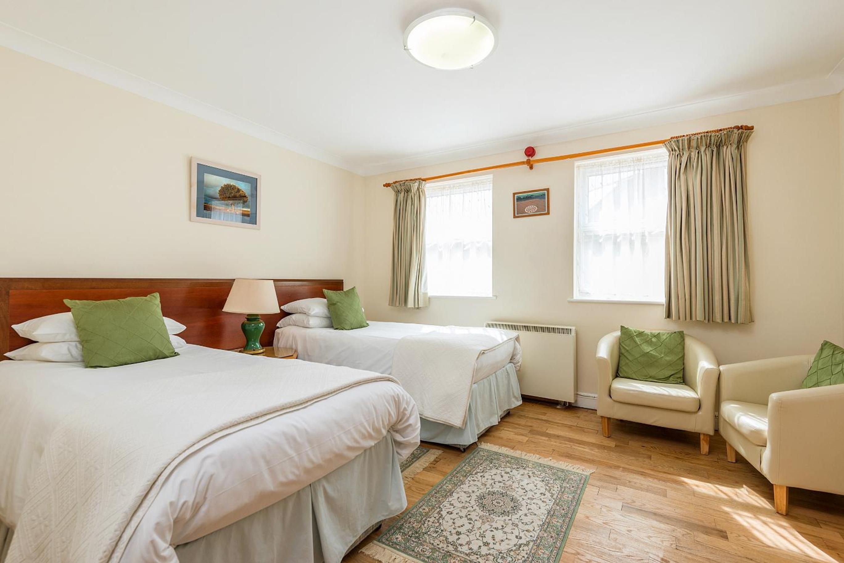 A hotel room with two beds, a sitting area with a chair, a radiator, and a window with curtains.