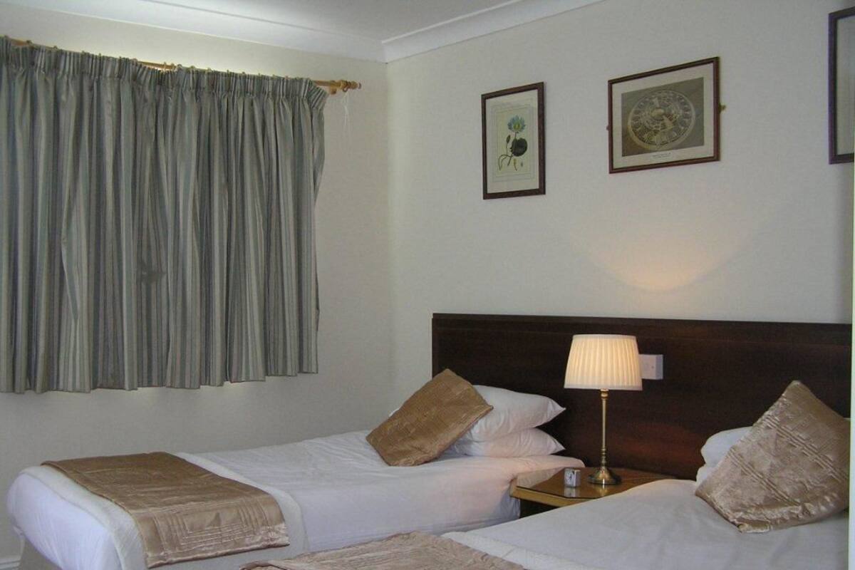 Habitación de hotel con dos camas individuales, cabecero de madera, mesita de noche con lámpara y cortina.
