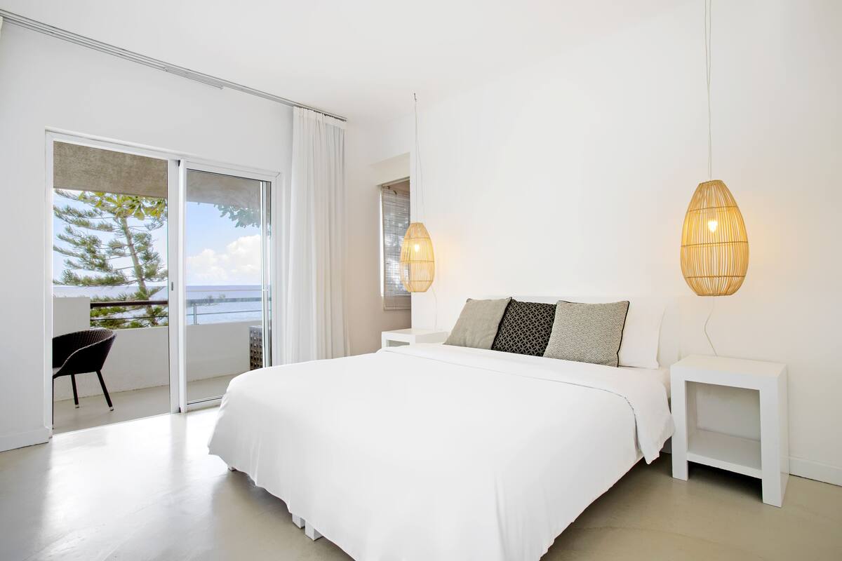 A bedroom with a large bed, white nightstands, and a view of the outdoors.