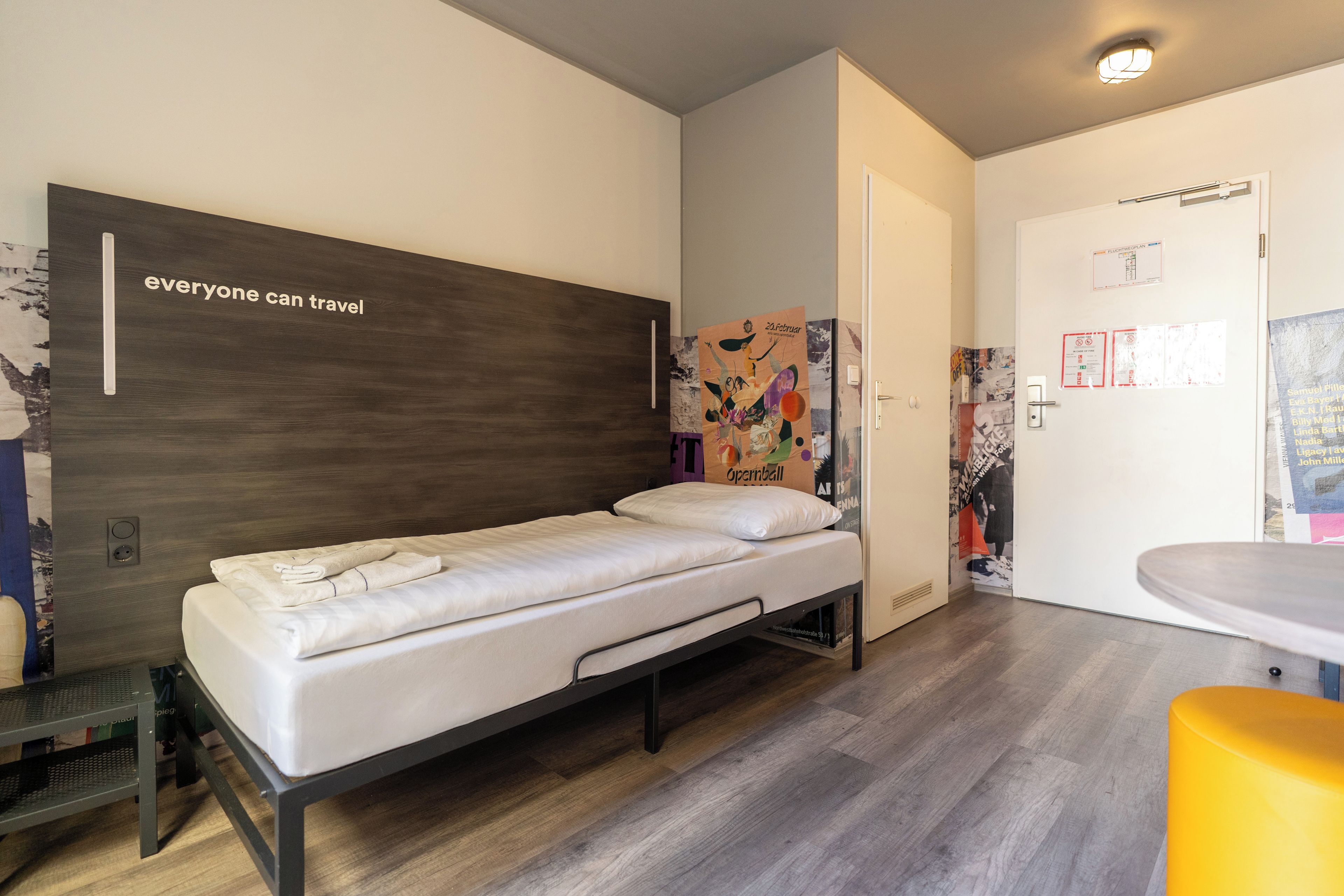 Un lit simple avec une tête de lit en bois, un petit bureau avec une chaise et une salle de bain équipée d’un lavabo et d’un miroir.