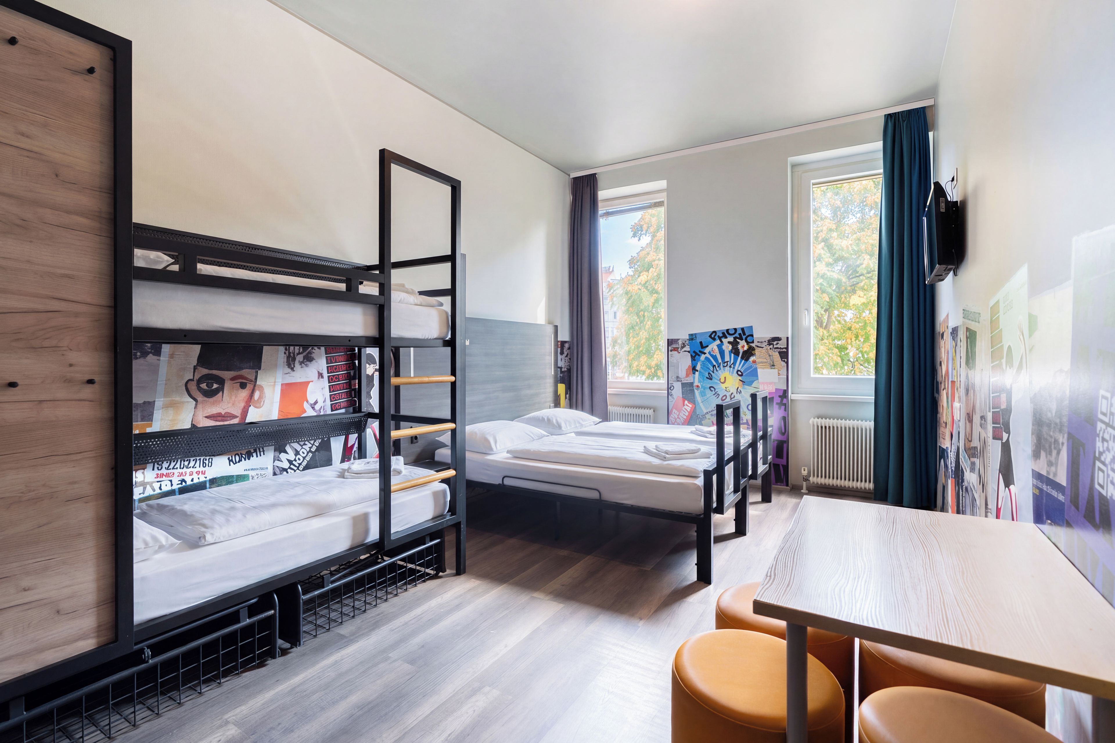A compact hotel room with bunk beds, a single bed, a small table, and a window with a view of trees.