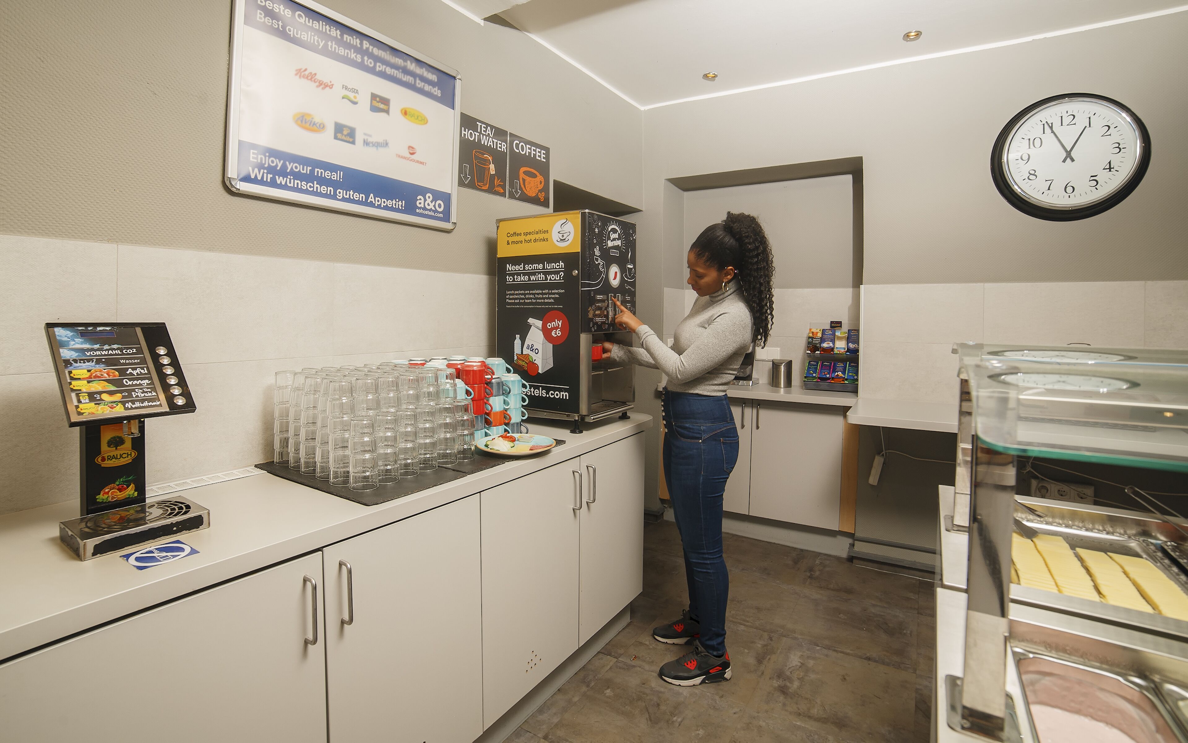 A woman at a coffee machine in a cafe with a menu board above and a clock on the wall.