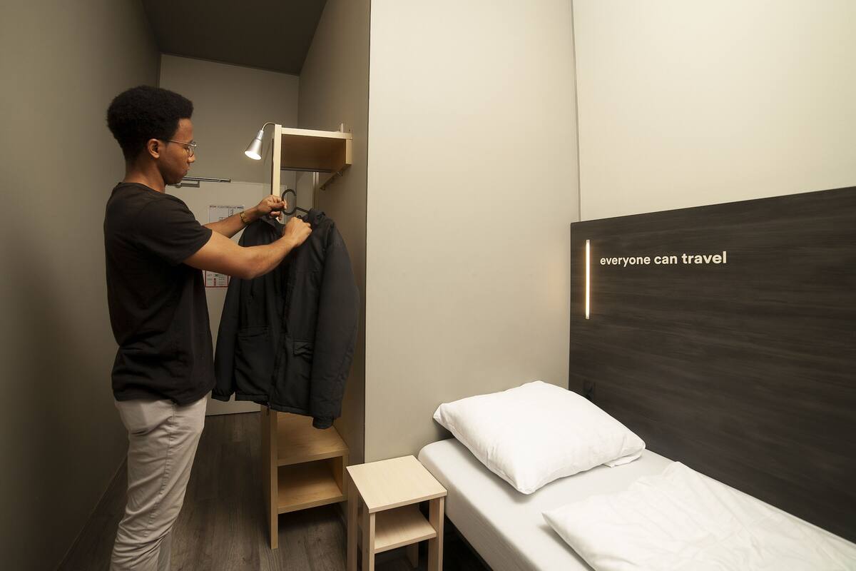 Ein Mann in schwarzem Hemd und beigen Hosen hängt Kleidung auf ein Holzgestell in einem Zimmer mit Bett und Wandregal.