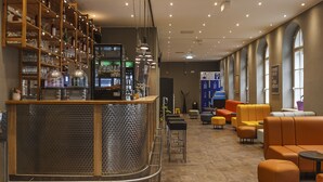 A bar area with shelves of bottles and glasses, a counter with a cash register, and seating with orange and gray cushions. - a&o Wien Stadthalle (Vienna)