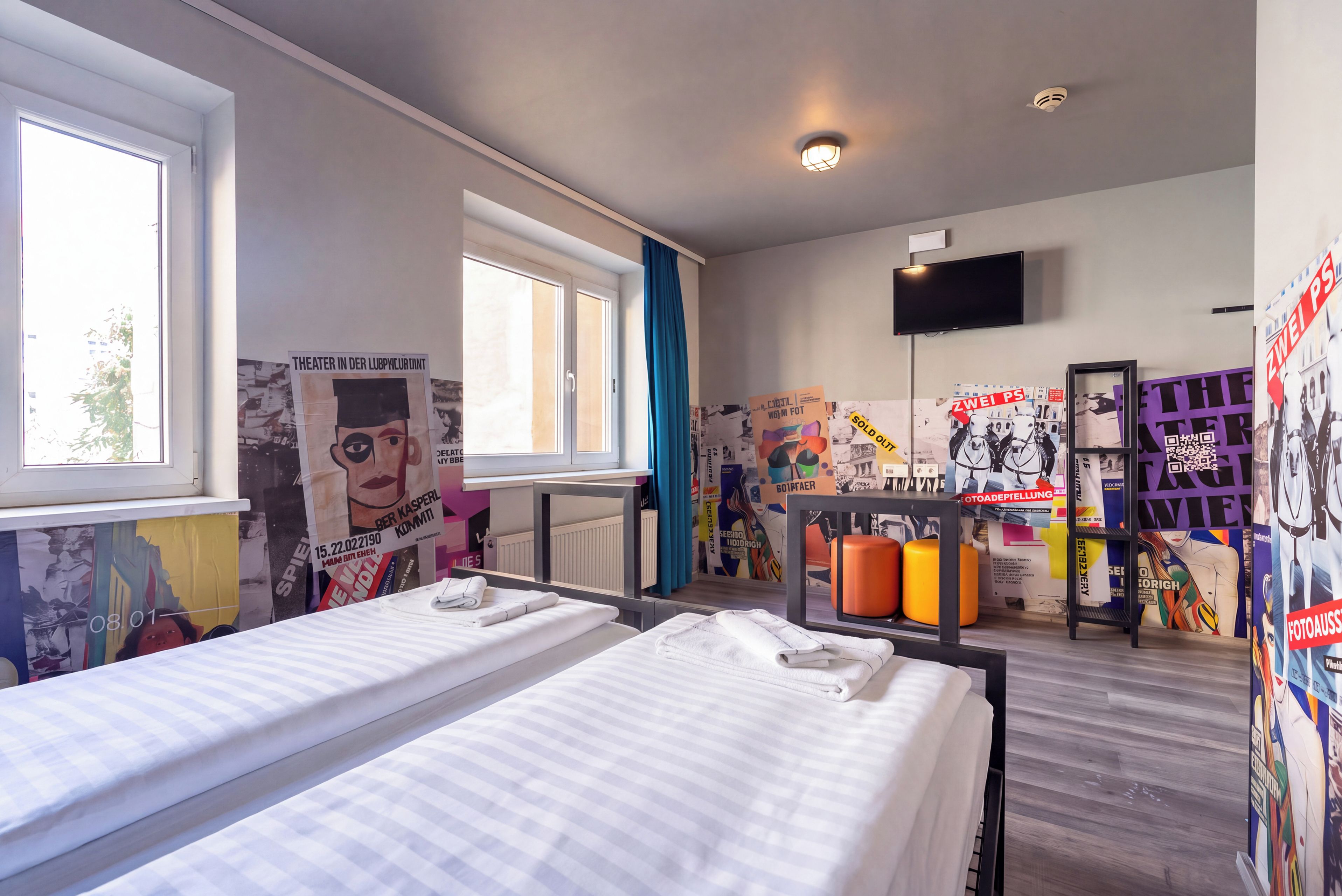 Une chambre d’hôtel avec deux lits, une fenêtre, un téléviseur et un mur orné de divers affiches, ainsi qu’une étagère avec différents objets.