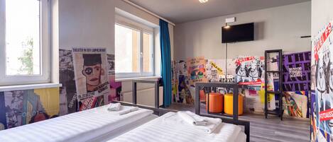 Une chambre d’hôtel avec deux lits, une fenêtre, un téléviseur et un mur orné de divers affiches, ainsi qu’une étagère avec différents objets.