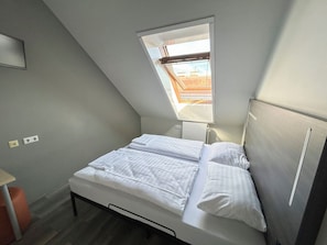Quarto pequeno e moderno com claraboia, cama com roupa de cama branca e radiador.