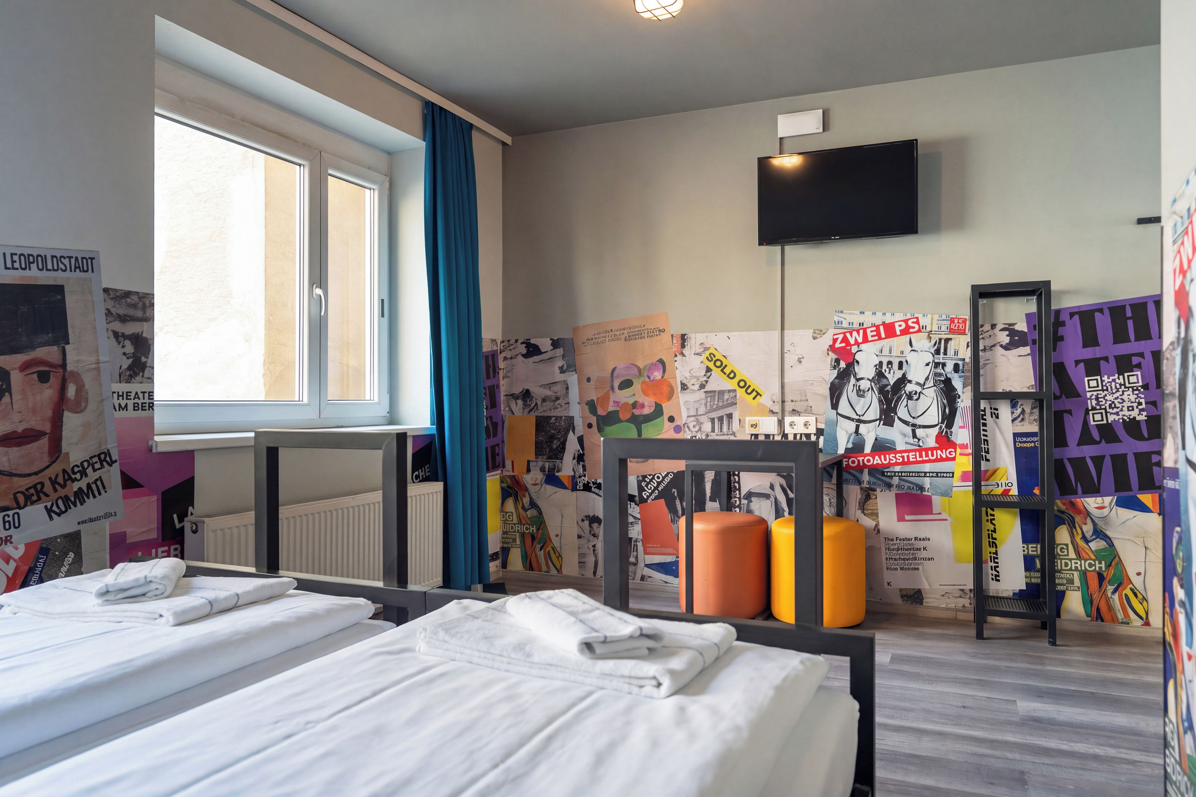 A hotel room with a bed, a window, a TV, and a wall decorated with various posters.