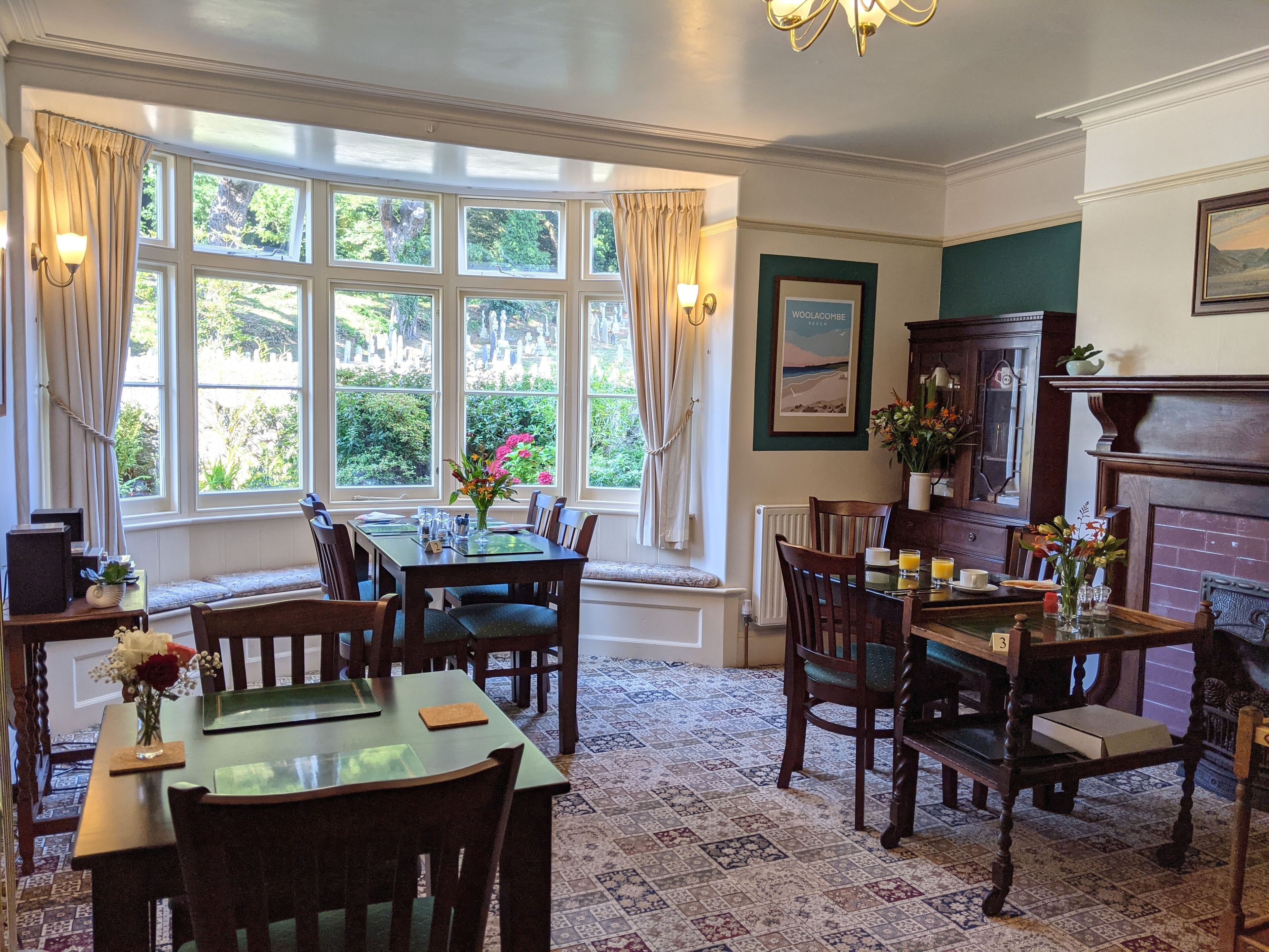 A dining area with a table set for four, surrounded by chairs. There are large windows with curtains, a fireplace, and a framed picture on the wall.
