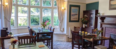 A dining area with a table set for four, surrounded by chairs. There are large windows with curtains, a fireplace, and a framed picture on the wall.