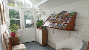 A cozy reading nook with a white wicker chair, a wooden bench, and a small bookshelf with books. - The Denes Guest House (Lynton)