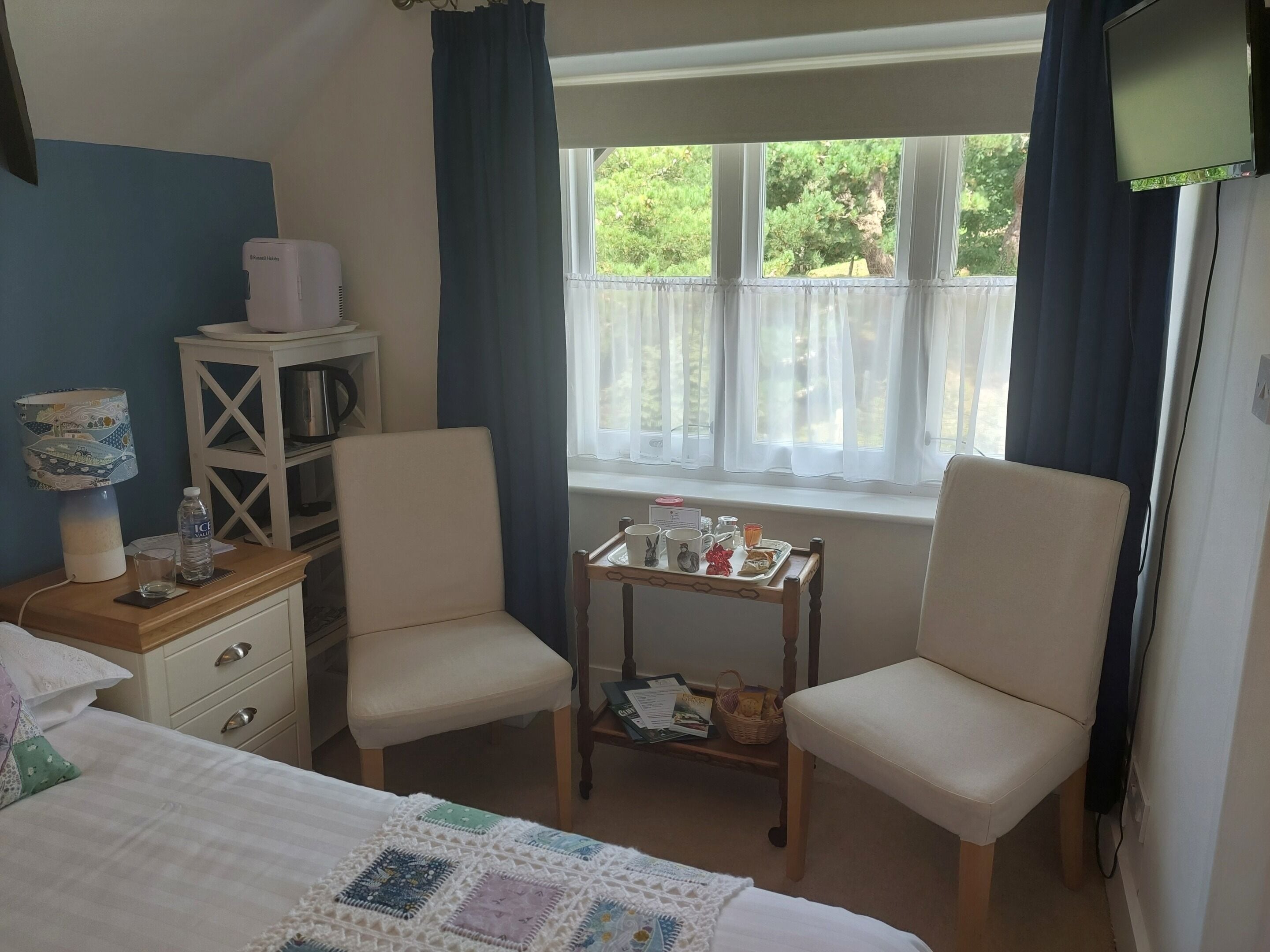 Una habitació d'hotel amb un llit, dues cadires, una taula petita amb una safata d'objectes, una finestra amb cortines, una estant amb un búllet i una làmpada.
