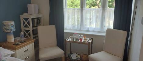 Una habitació d'hotel amb un llit, dues cadires, una taula petita amb una safata d'objectes, una finestra amb cortines, una estant amb un búllet i una làmpada.
