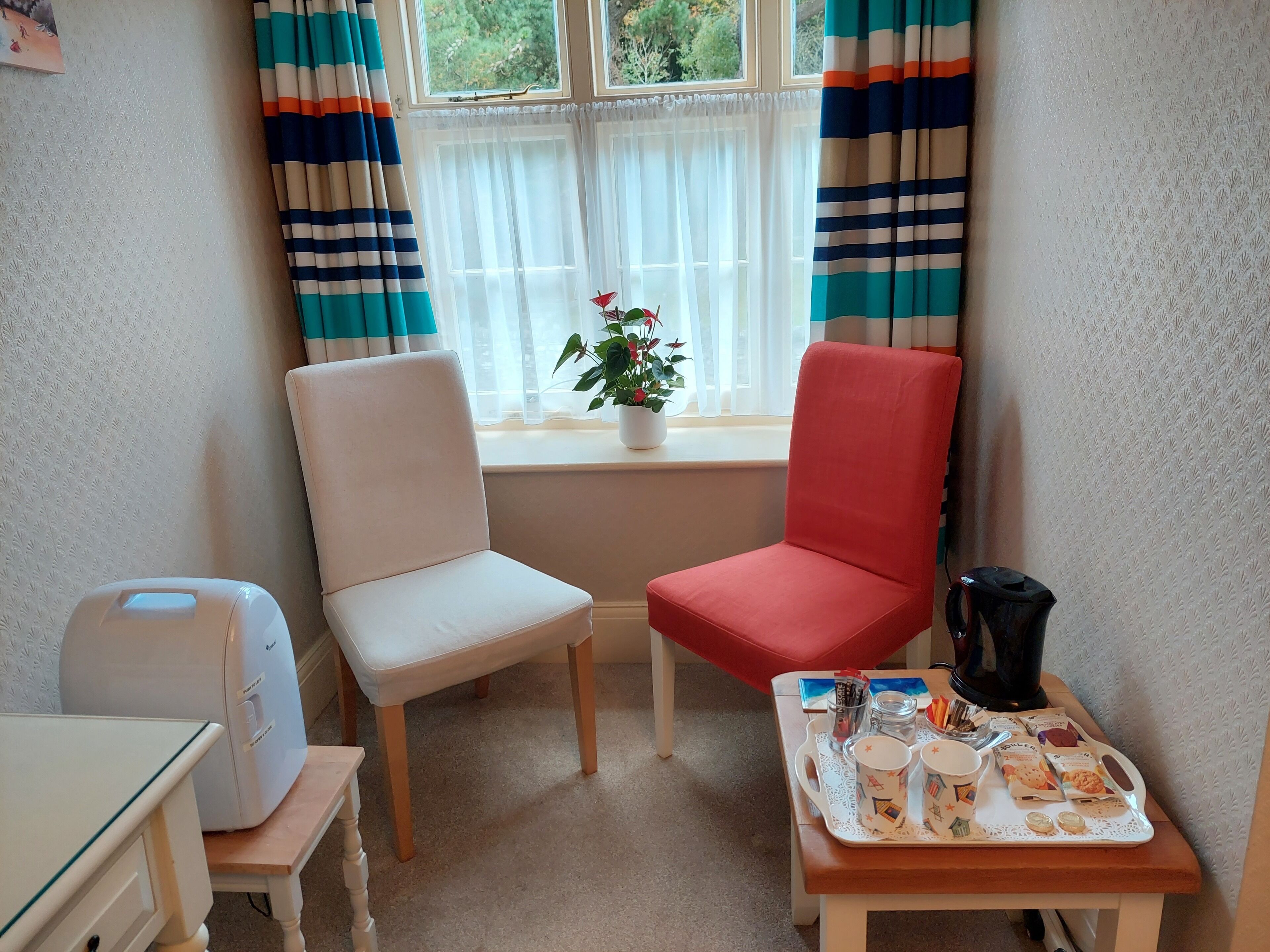 Een hoekzaal met twee stoelen, een tafeltje met een dienblad en spullen, een raam met gordijnen, en een kamerplant.