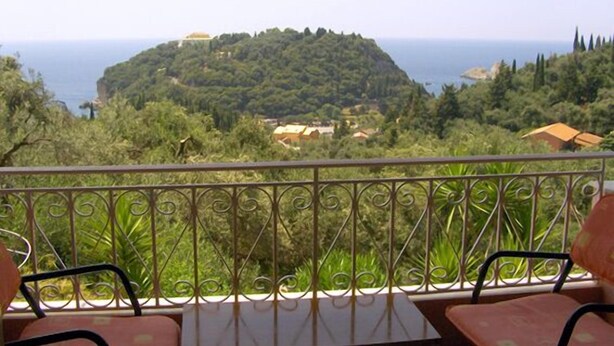 Un balcon doté de balustrades en métal et de deux chaises, offrant une vue sur un paysage verdoyant et luxuriant, avec une colline et la mer en arrière-plan.