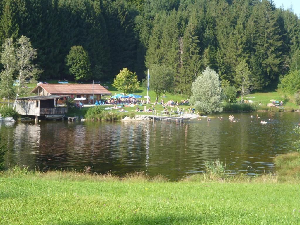 Eine Seenlandschaft mit einem hölzernen Steg, einem kleinen Gebäude und Menschen, die am Wasser entspannen.