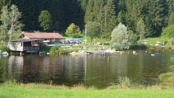 Eine Seenlandschaft mit einem hölzernen Steg, einem kleinen Gebäude und Menschen, die am Wasser entspannen.