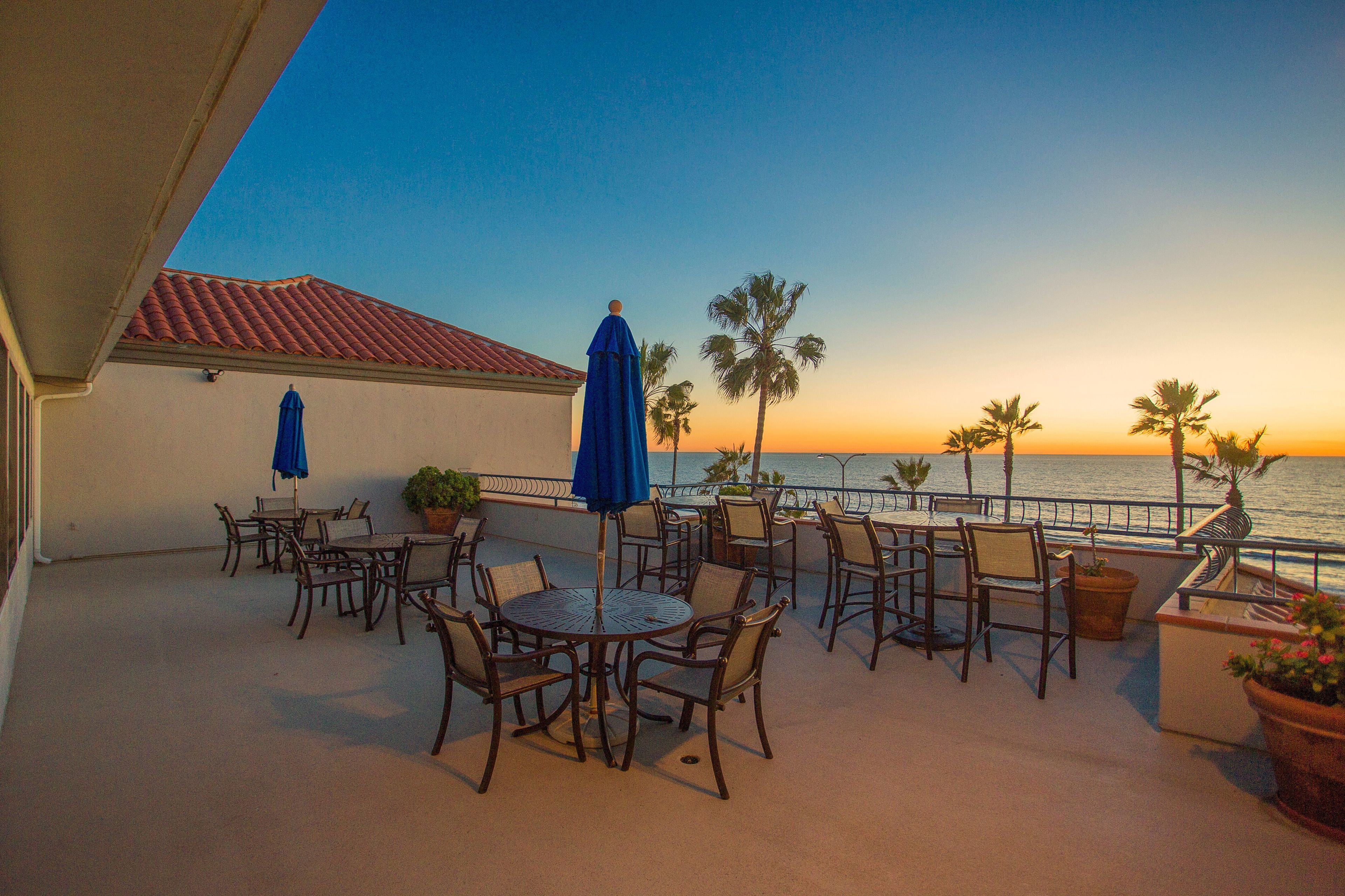 An outdoor patio with tables and chairs, a blue umbrella, and a view of the ocean at sunset.